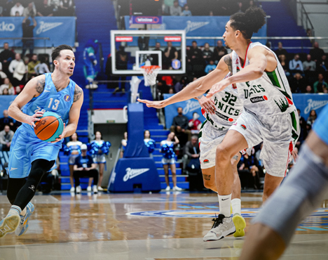 На изображении два баскетболиста: один в голубой форме с мячом, другой в серой форме защищается. Сзади видны зрители на трибунах. Игроки находятся на паркете в спортивном зале