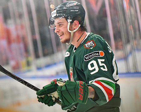 Хоккеист в форме зелёного цвета с номером 95 на рукаве стоит на льду с клюшкой в руке. На шлеме и форме размещены логотипы команды и спонсоров. Он улыбается, фон размыт