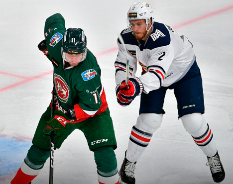 На изображении два хоккеиста в игровой борьбе за шайбу на льду. Спортсмен в зелёной форме и игрок в бело-синей форме активно взаимодействуют, находясь в движении. На заднем плане видна разметка хоккейной площадки