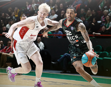 На изображении два баскетболиста в игровом эпизоде на площадке. Игрок в белой форме защищается, пытаясь остановить соперника в тёмной форме с мячом. На заднем плане видно зрителей